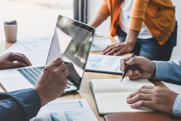 Three people work at a table with graphs, a laptop displaying charts, and a notebook, discussing data and writing notes.