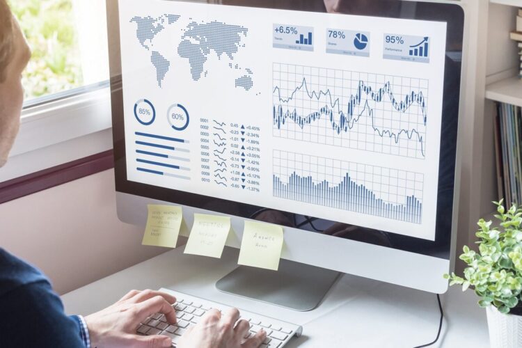 Person analyzing charts and graphs on a computer screen, displaying global statistics, line graphs, bar charts, and data visualizations; sticky notes are attached to the monitor.