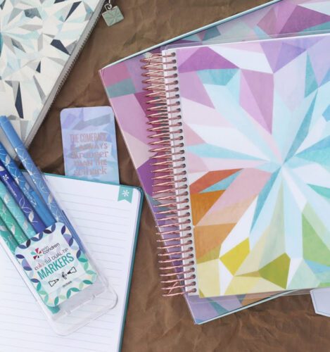 Assorted stationery items including a spiral notebook with a geometric pastel cover, pens, a zip pouch, and a card on a brown surface.