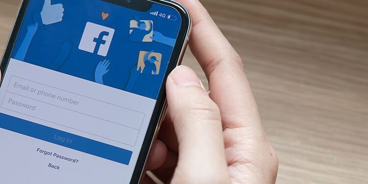 Person holding a smartphone with the Facebook login screen displayed, showing fields for email or phone number and password, on a wooden desk.