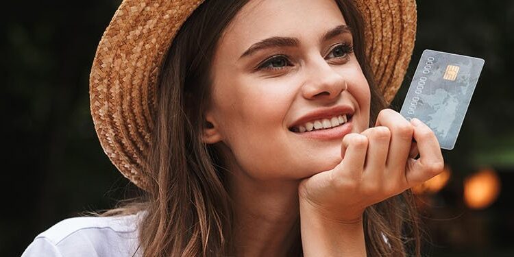 A woman wearing a straw hat smiles while holding a credit card outdoors, with blurred greenery in the background.