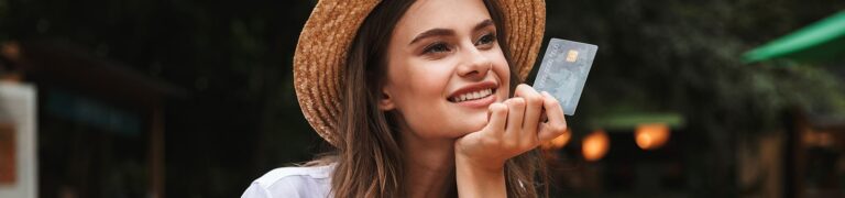 A woman wearing a straw hat smiles while holding a credit card outdoors, with blurred greenery in the background.