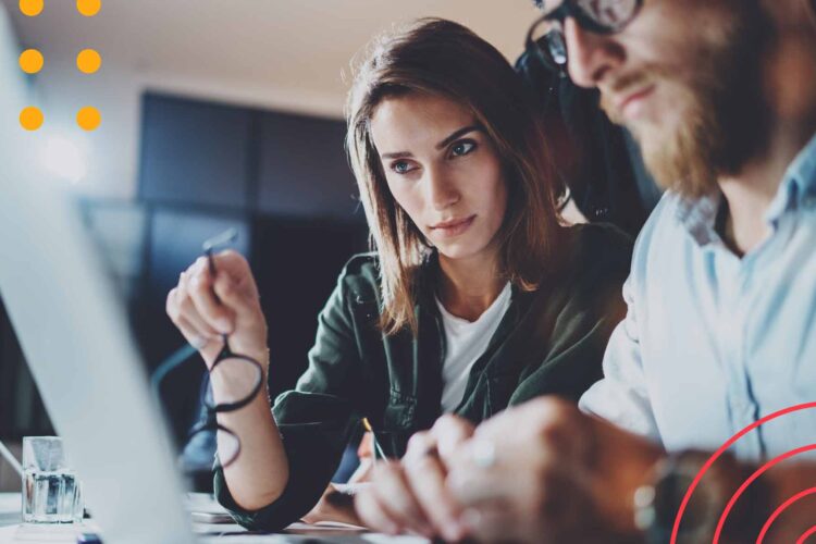 Two people sit at a desk looking intently at a laptop screen, with one holding glasses and the other typing.