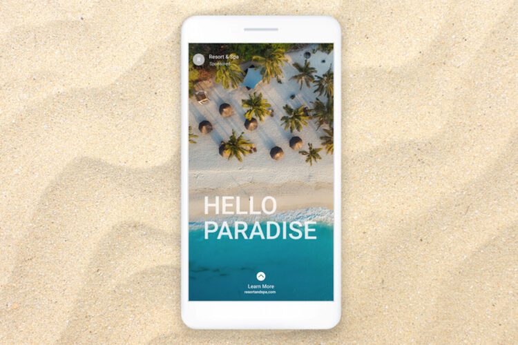 A smartphone on sand displays an aerial view of a tropical beach with palm trees, huts, and blue water. The screen reads "HELLO PARADISE" with a "Learn More" button below.