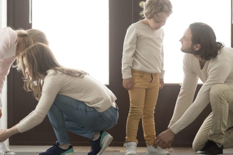 Two adults are kneeling to help two children put on their shoes in front of a door.