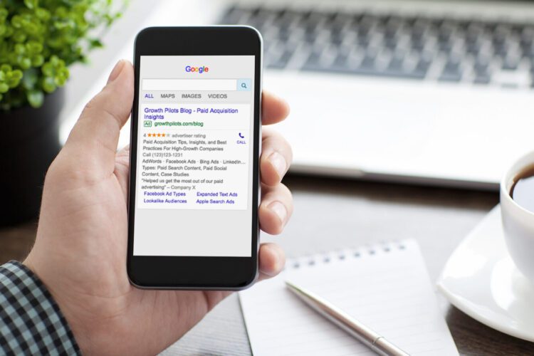 A person holds a smartphone displaying Google search results, next to a notepad, pen, and cup of coffee on a desk.