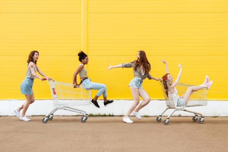 Four young women playfully ride and push two shopping carts outside against a yellow wall, appearing cheerful and energetic.