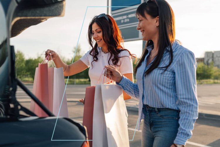 Two women standing by a car trunk placing shopping bags inside, outdoors on a sunny day.