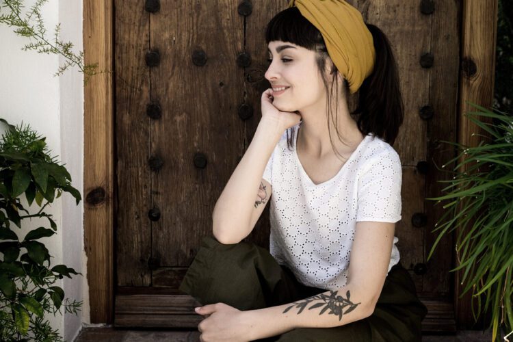 A woman with dark hair and a yellow headband sits in front of a wooden door, resting her chin on her hand, surrounded by green plants.