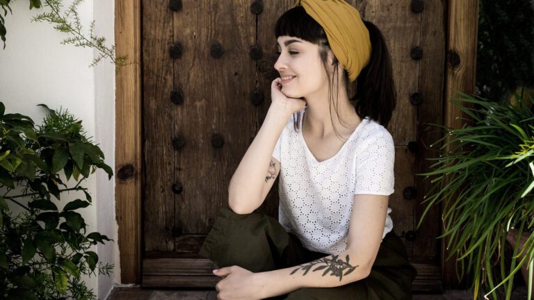 A woman with dark hair and a yellow headband sits in front of a wooden door, resting her chin on her hand, surrounded by green plants.
