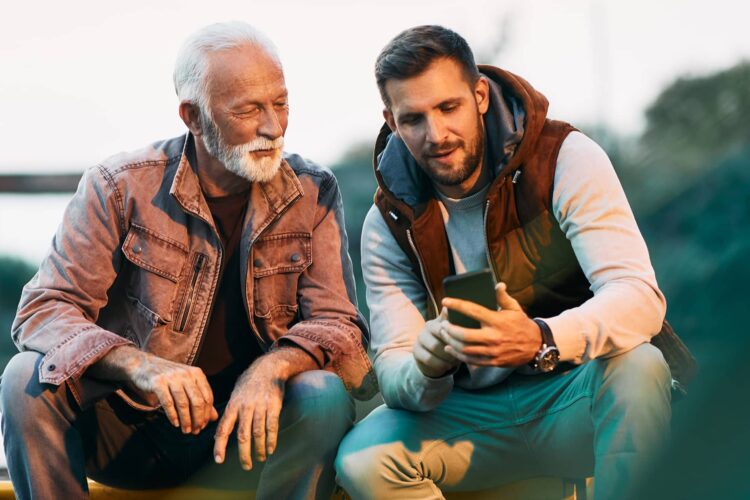 An older man and a younger man sit outdoors; the younger man shows something on his smartphone to the older man.