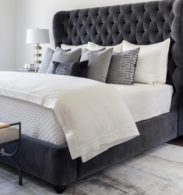 A modern bedroom features a tufted dark gray upholstered bed with white bedding, multiple decorative pillows, a bench at the foot, and a bedside table with a lamp.