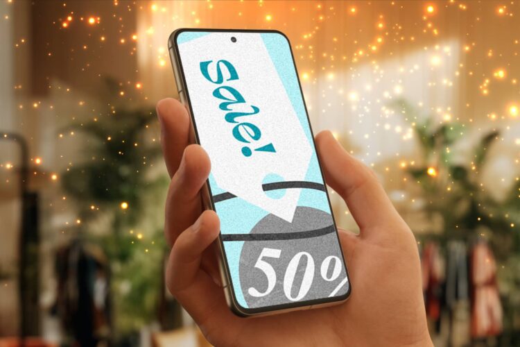A hand holds a smartphone displaying a "Sale! 50%" sign, with blurred indoor greenery and sparkling lights in the background.