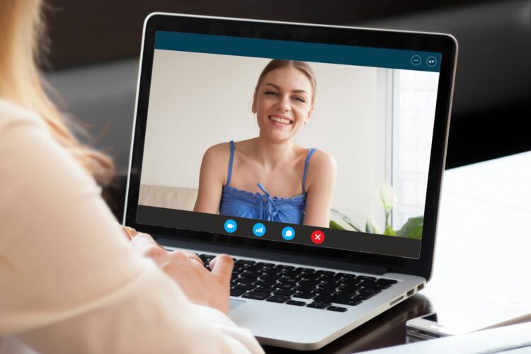 A person participates in a video call on a laptop, viewing a woman in a blue top who is smiling at the camera.