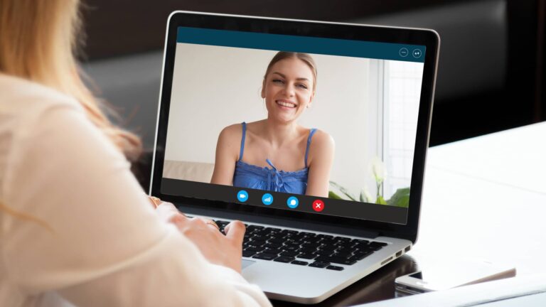 A person participates in a video call on a laptop, viewing a woman in a blue top who is smiling at the camera.