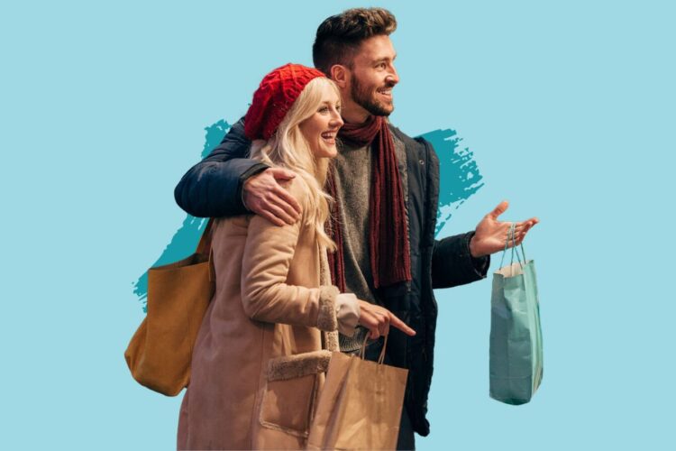 A smiling man and woman wearing winter clothes hold shopping bags and stand together against a light blue background.