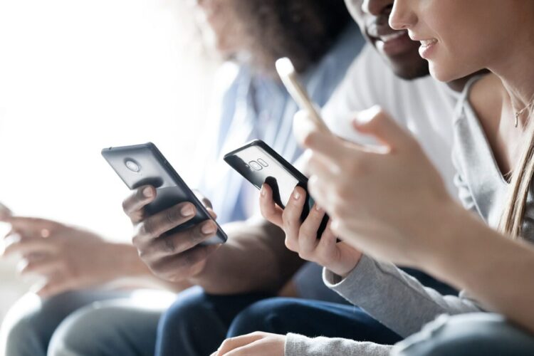 Three people sitting close together, holding and using smartphones, with their faces partially visible and focused on their screens.