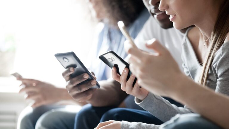Three people sitting close together, holding and using smartphones, with their faces partially visible and focused on their screens.