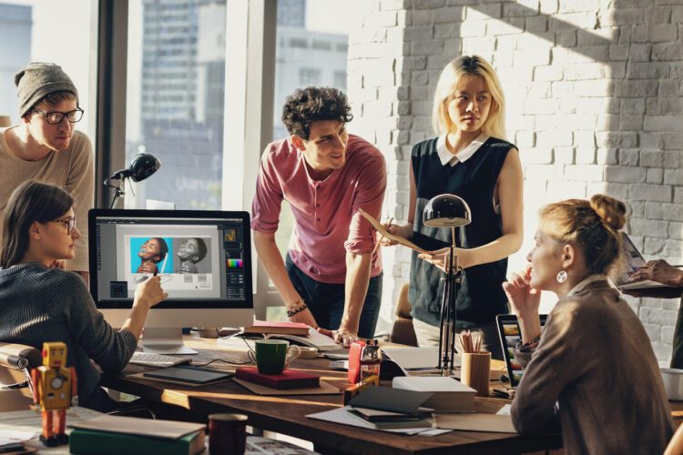 A group of six people collaborates around a desk in a modern office, with one computer displaying a digital portrait on the screen.