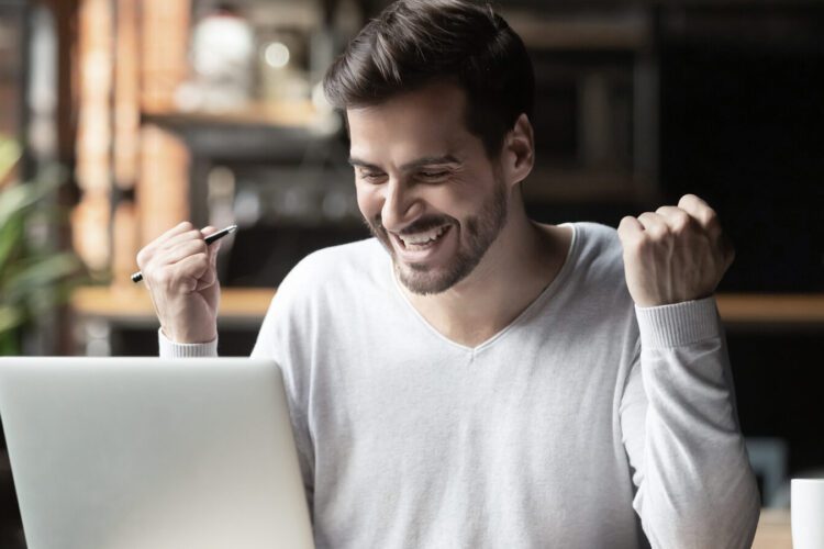 A man sits at a desk in front of a laptop, smiling and raising his fists in excitement while holding a pen, with a mug and plant in the background.