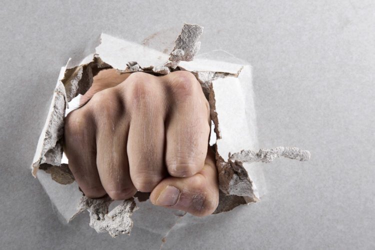 A clenched fist punches through and breaks a sheet of white drywall, creating a hole with loose debris around the edges.