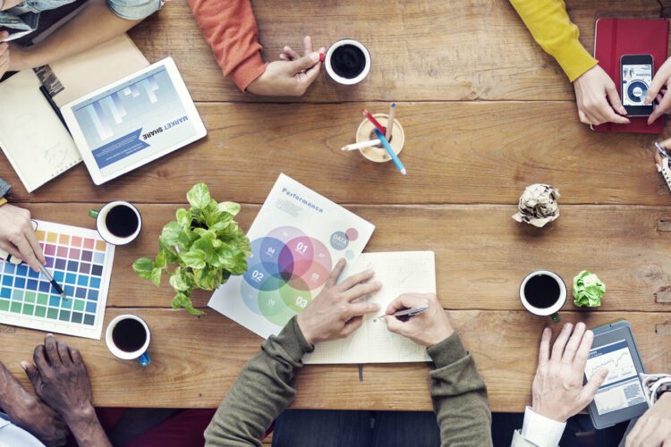 A group of people collaborate at a wooden table with laptops, charts, color swatches, coffee cups, and notepads visible.