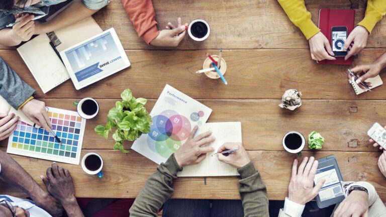 A group of people collaborate at a wooden table with laptops, charts, color swatches, coffee cups, and notepads visible.
