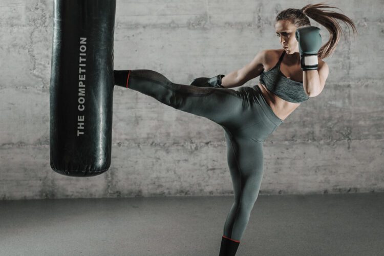 A woman in athletic wear performs a high kick against a punching bag in a gym setting.