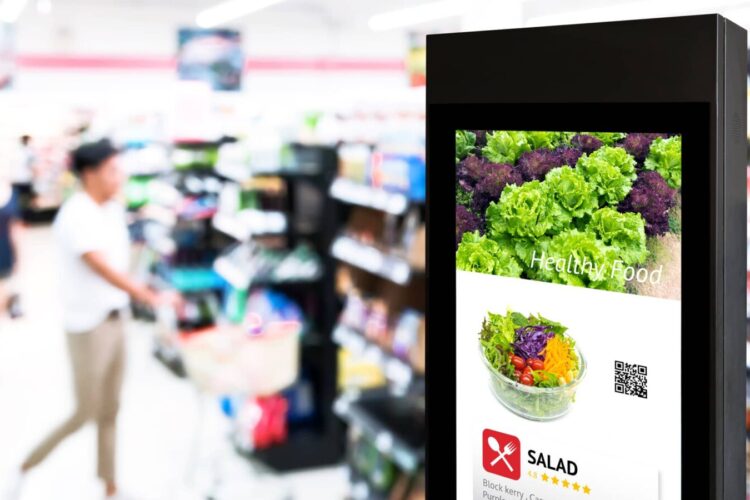 A digital kiosk in a grocery store displays an image of a salad with the words “Healthy Food”; blurred shoppers and store shelves are visible in the background.