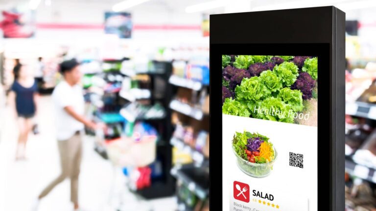 A digital kiosk in a grocery store displays an image of a salad with the words “Healthy Food”; blurred shoppers and store shelves are visible in the background.