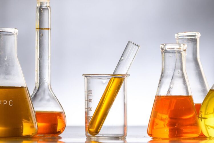Five laboratory glassware pieces containing orange liquid, including a beaker with a test tube inside, are arranged on a reflective surface against a neutral background.