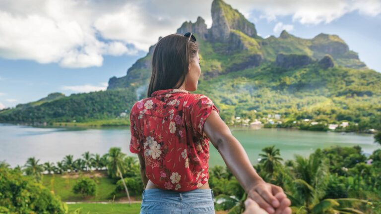 A woman in a red floral shirt holds someone's hand and looks at a mountainous tropical landscape with water and greenery.