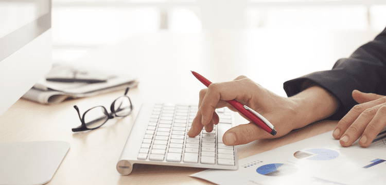 A person uses a computer keyboard while holding a red pen and reviewing printed graphs and charts on a desk. Glasses are placed nearby.