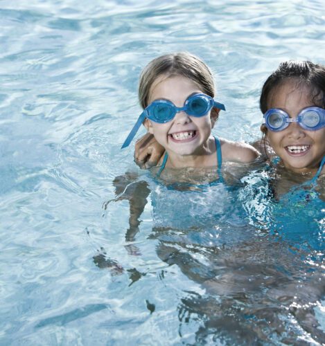 Two children wearing blue swim goggles smile while standing together in a swimming pool.