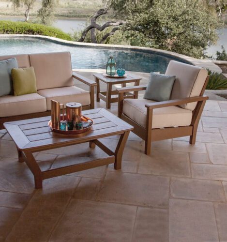Outdoor patio with beige cushioned seating, wooden tables, and decorative items beside a pool overlooking a natural landscape.
