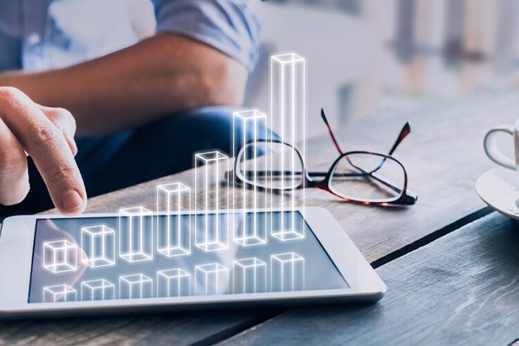 A person uses a tablet displaying a digital bar graph, with eyeglasses and a cup on a wooden table.