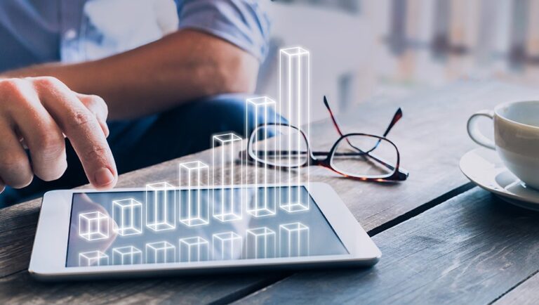 A person uses a tablet displaying a digital bar graph, with eyeglasses and a cup on a wooden table.