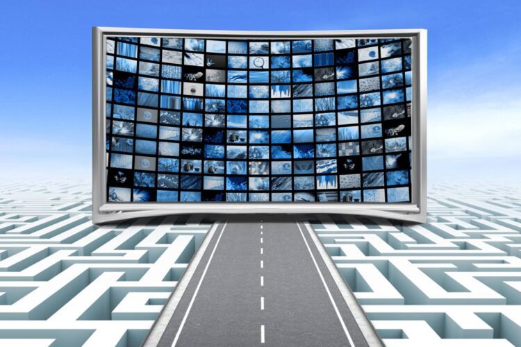 A curved screen displaying multiple blue-toned images stands at the end of a road passing over a maze, with a blue sky in the background.