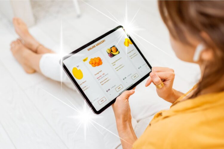 A person sitting on the floor uses a tablet to shop for groceries online, with fruit options displayed on the screen.