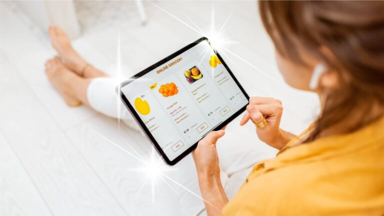 A person sitting on the floor uses a tablet to shop for groceries online, with fruit options displayed on the screen.