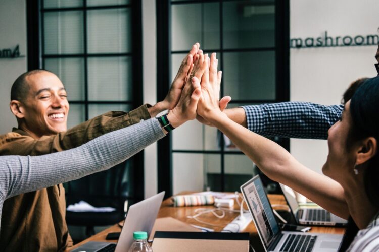 Four people sit at a table with laptops, reaching in to give a group high-five, appearing engaged in a collaborative work environment.