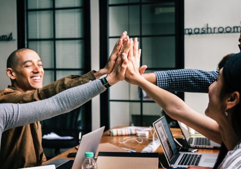 Four people sit at a table with laptops, reaching in to give a group high-five, appearing engaged in a collaborative work environment.