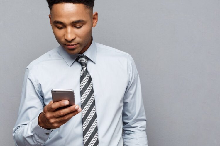 A man in a dress shirt and striped tie looks down at a smartphone against a plain gray background.