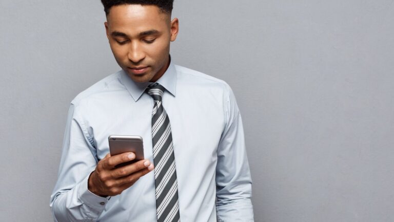 A man in a dress shirt and striped tie looks down at a smartphone against a plain gray background.