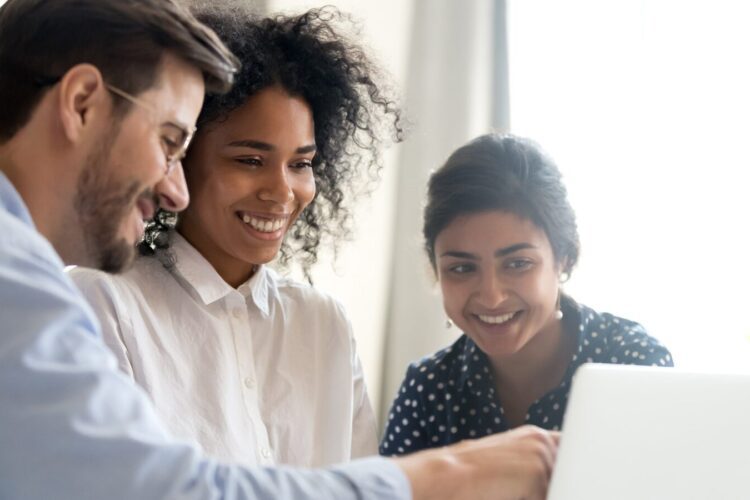 Three people sit together at a table, smiling as they look at a laptop screen in a bright indoor setting.