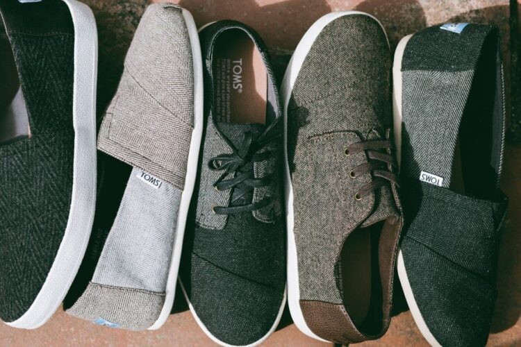 Five pairs of TOMS casual shoes in various neutral colors are arranged side by side on a stone surface.