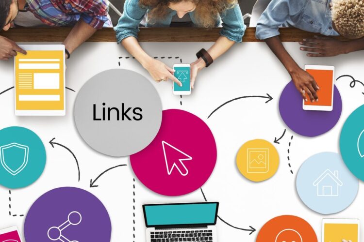 Three people sit at a table covered with colorful diagrams and icons related to digital links, using a laptop, tablet, and documents.