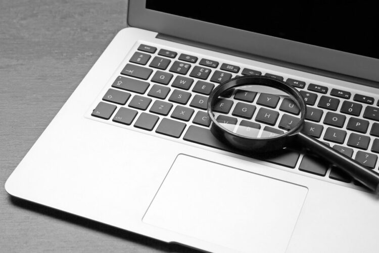 A magnifying glass rests on the keyboard of an open laptop on a wooden surface.