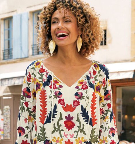 Woman with curly hair smiles outdoors, wearing a cream-colored top with colorful floral embroidery and large gold leaf-shaped earrings.