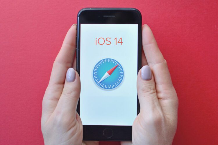 A person holds an iPhone displaying the Safari browser icon and “iOS 14” text on a white screen, against a solid red background.